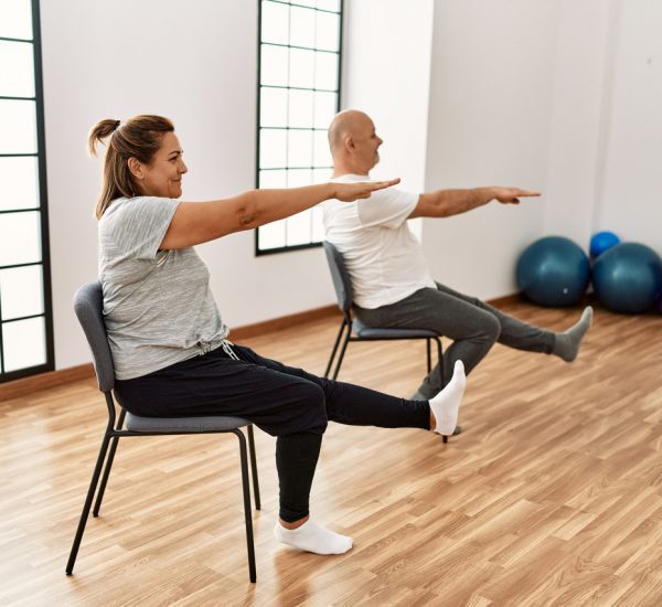 Middle age hispanic couple stretching using chair at sport center.